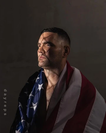 Adult male draped with an American flag looking off-camera in a low-key indoor portrait with hard rim lighting against a grey background.