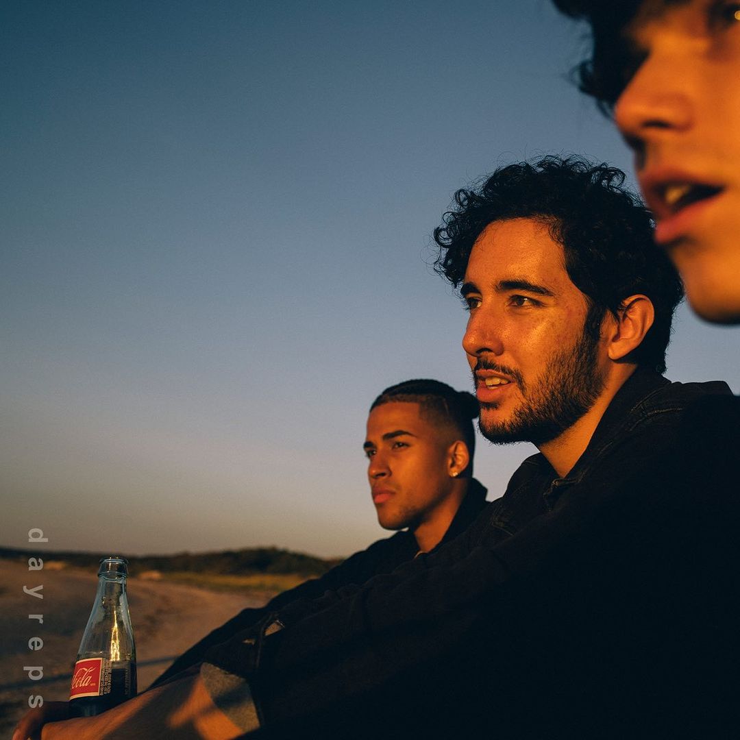Three young men are silhouetted by a bright sky as they sit near a bottle on a beach. Golden hour lighting creates a warm atmosphere.