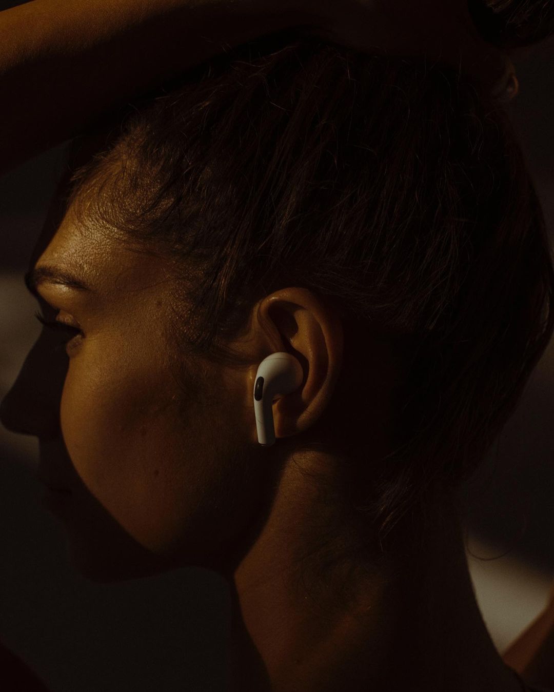 Profile shot of an adult female's face with an earbud in her ear, warm and dark lighting, and a neutral expression.