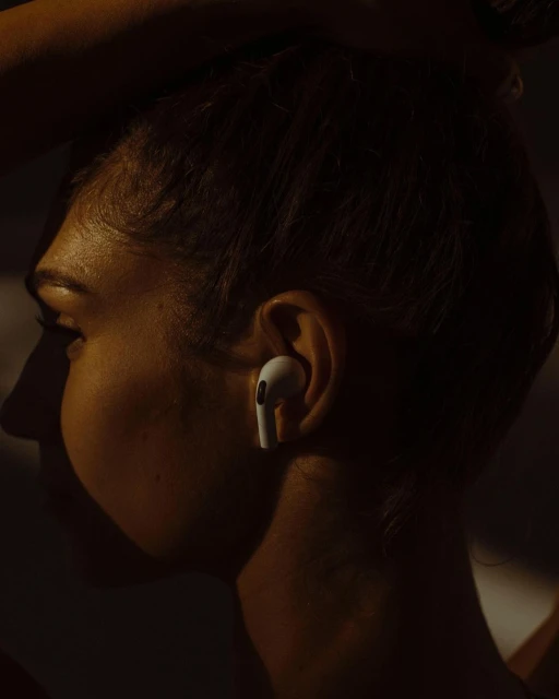 Profile shot of an adult female's face with an earbud in her ear, warm and dark lighting, and a neutral expression.