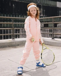 Child model wearing a pink tracksuit and headband holding a tennis racket on a rooftop by Nick Laham