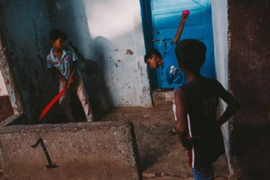 Three young boys playing street cricket in a narrow alleyway with a blue door and a painted number one on a concrete wall.