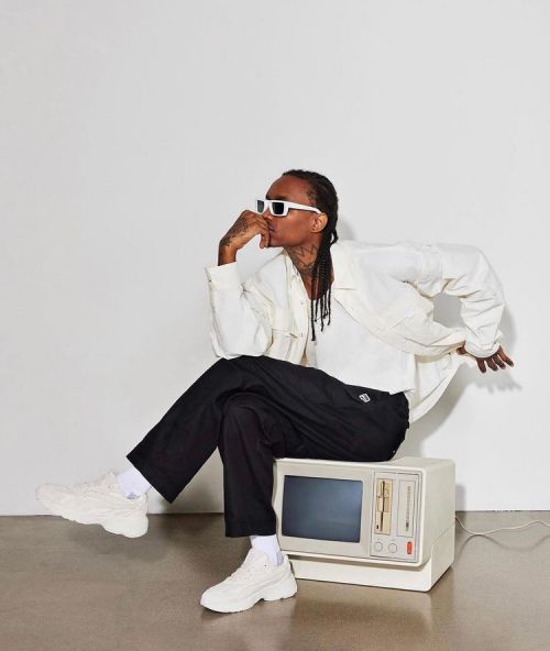 A man in white sunglasses and a light jacket poses, sitting on a vintage television in a minimalist studio setting with neutral tones.