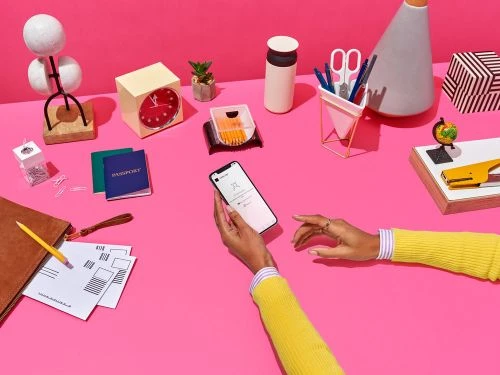 Woman's hands holding smartphone on a pink surface amid desk accessories, including a clock, passport, office supplies, and decor.