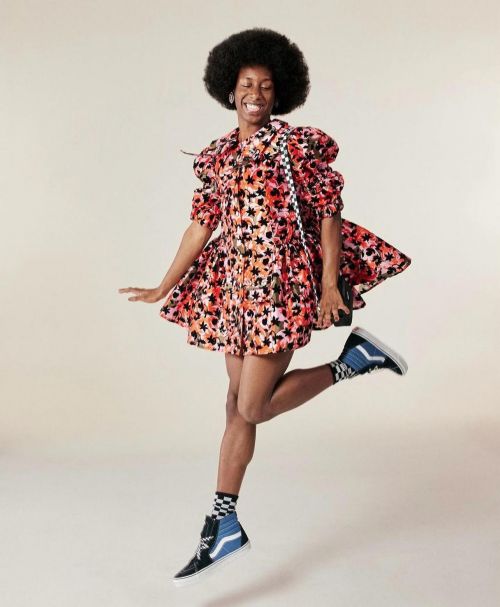 A happy Black woman with afro hairstyle jumps with joy wearing a vibrant floral-patterned mini dress and Vans sneakers in a studio shot.