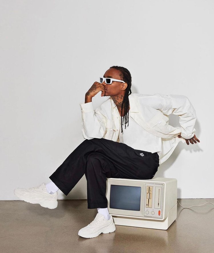 Stylish adult male posing while seated on a retro television set indoors, wearing white sunglasses, jacket, and chunky sneakers.