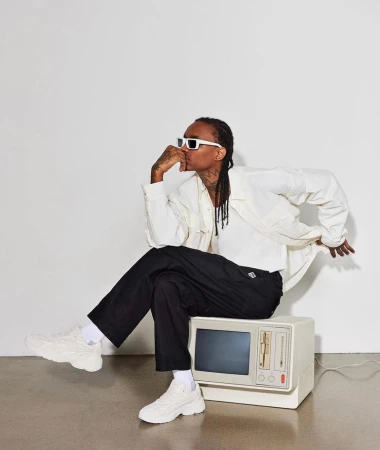 Stylish adult male posing while seated on a retro television set indoors, wearing white sunglasses, jacket, and chunky sneakers.