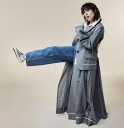 Woman in grey coat and blue jeans kicking her leg out, wearing checkered sneakers against a plain off-white background