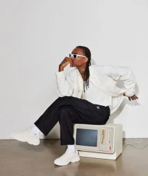 Man wearing white sunglasses and oversized shirt seated on a vintage computer against a white wall.