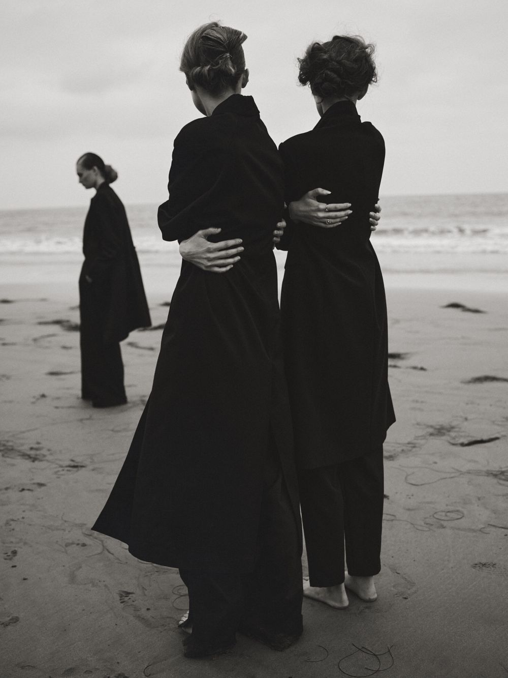 Three women stand on a beach, backs to the camera, in long black coats. Muted tones, soft light, and ocean background.