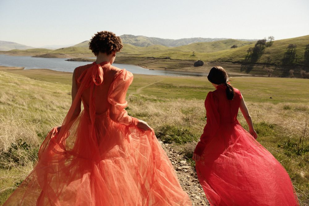 Two adult females wearing red and orange tulle dresses walk on a grassy hill near a lake on a sunny day; fashion editorial photography.