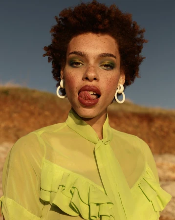 Close-up portrait of a woman with short curly brown hair wearing a lime green high-neck blouse and white hoop earrings under golden hour sunlight.
