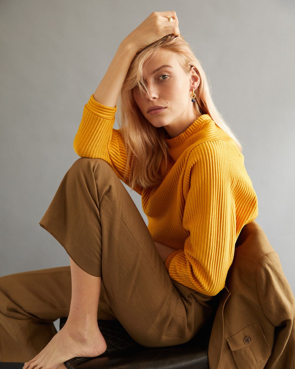 A blonde woman poses indoors wearing a yellow sweater, brown pants, and earrings, seated with a jacket draped on a chair. 