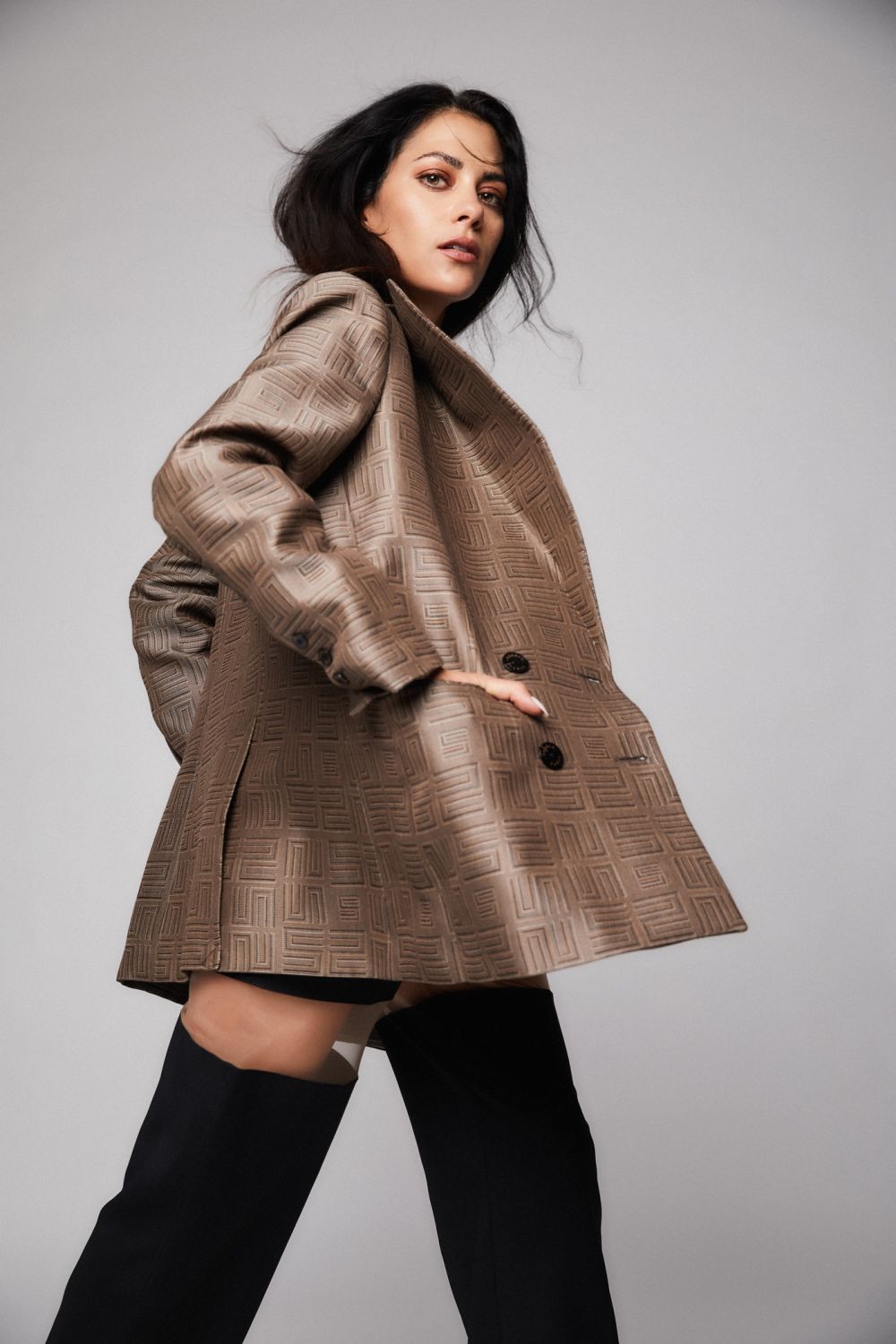 Fashionable woman in oversized patterned blazer and thigh-high boots poses in a studio setting against a neutral gray background.
