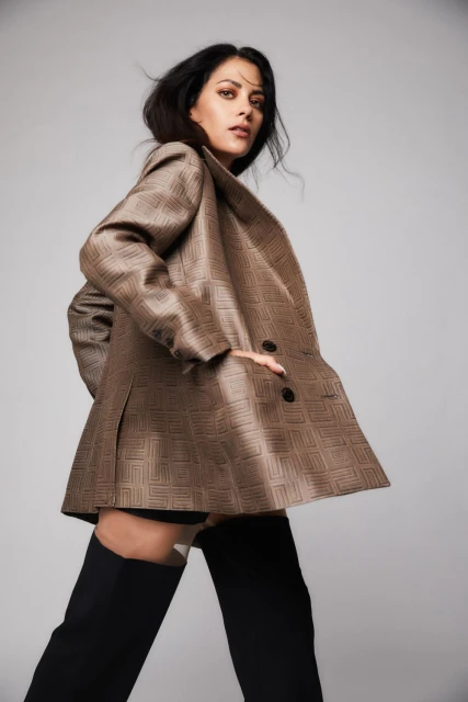 Fashionable woman in oversized patterned blazer and thigh-high boots poses in a studio setting against a neutral gray background.