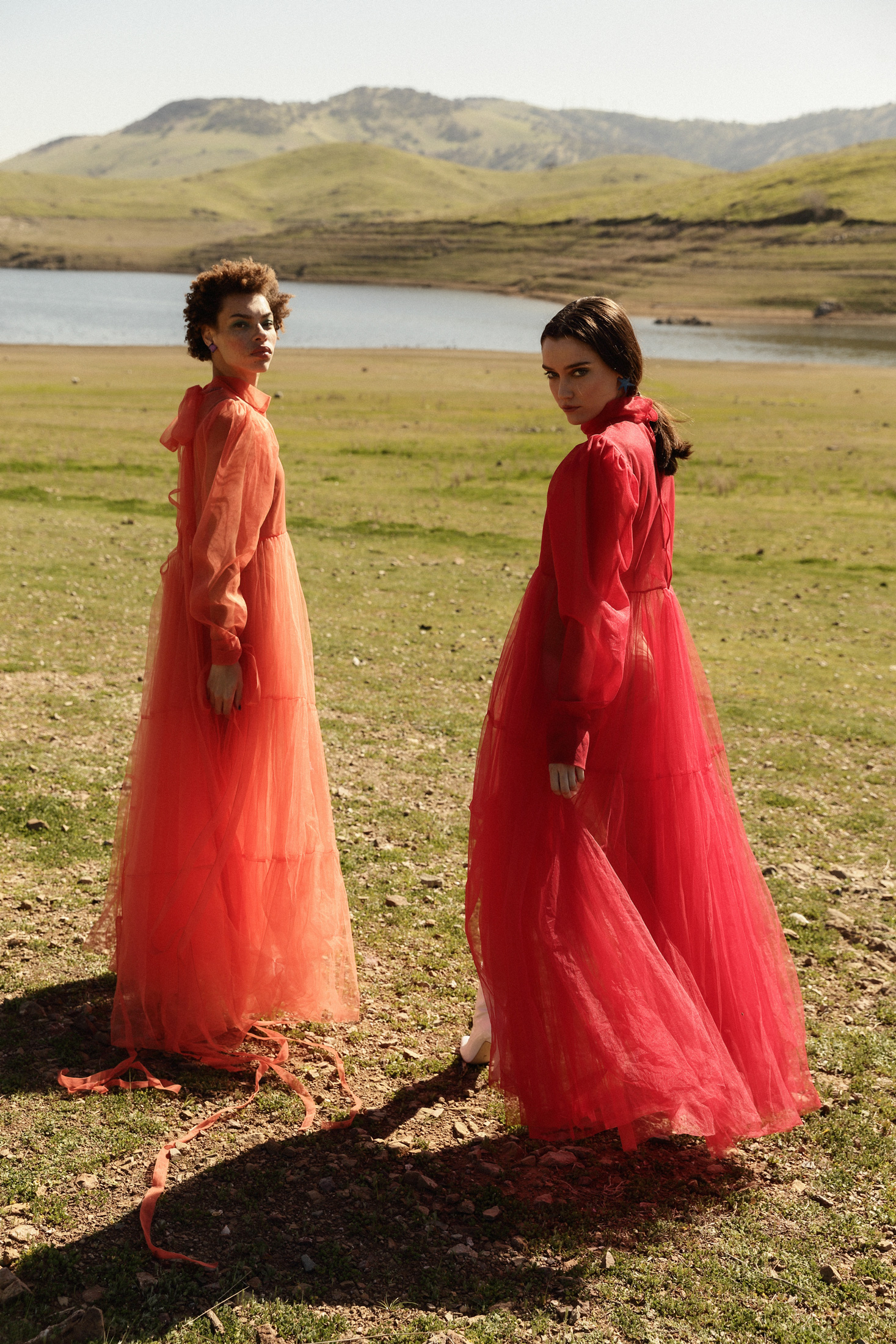 Two women in orange and red sheer dresses pose in a grassy field with a lake and mountains in the background. Soft sunlight.