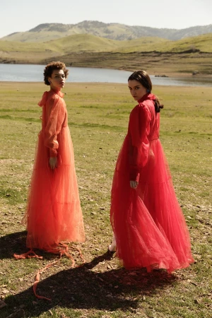 Two women in orange and red sheer dresses pose in a grassy field with a lake and mountains in the background. Soft sunlight.