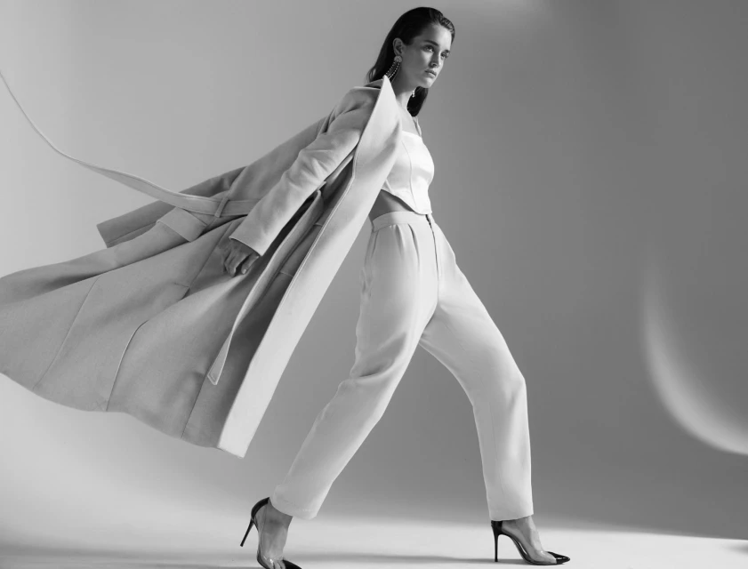 Elegant woman strides in a light-colored pantsuit and coat with heels, against a simple studio backdrop in a fashion forward pose.