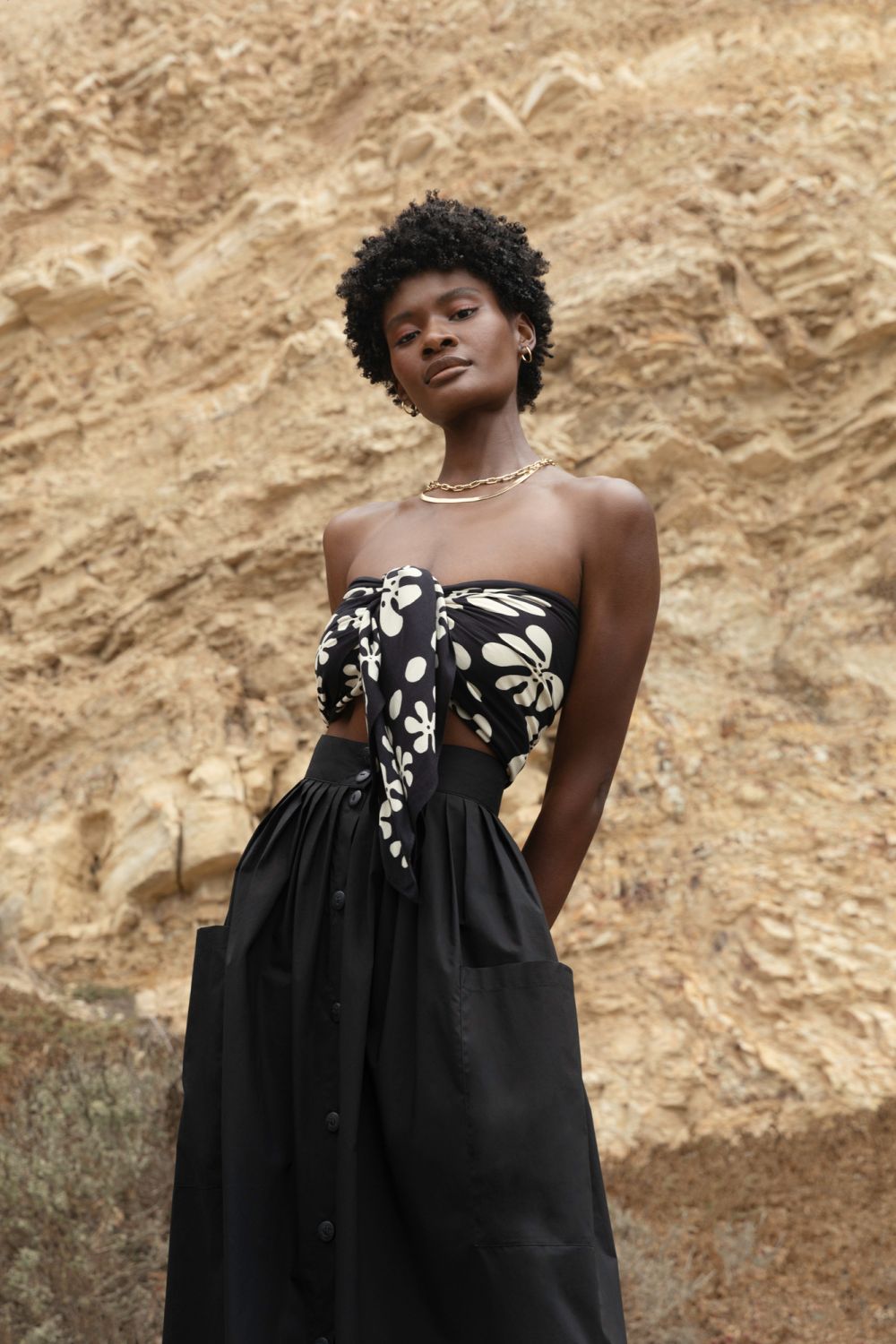 Adult Black woman posing in front of tan rock formation wearing a flower-patterned halter top and a long black skirt with visible pockets.