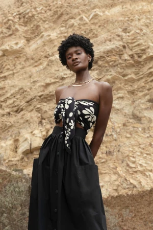 Adult Black woman posing in front of tan rock formation wearing a flower-patterned halter top and a long black skirt with visible pockets.