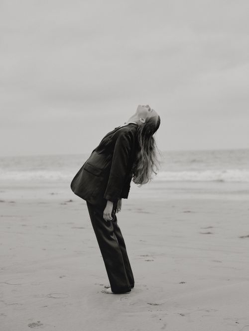 Woman in a black suit leaning backward on a sandy beach under an overcast sky, styled by Karlee Parrish.