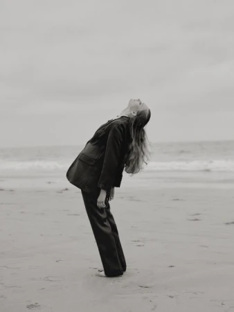 Woman in a black suit leaning backward on a sandy beach under an overcast sky, styled by Karlee Parrish.