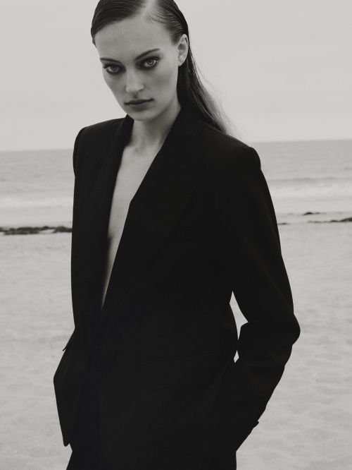 Monochrome portrait of a woman in a black blazer posing on a beach. She gazes at the camera with a neutral expression.