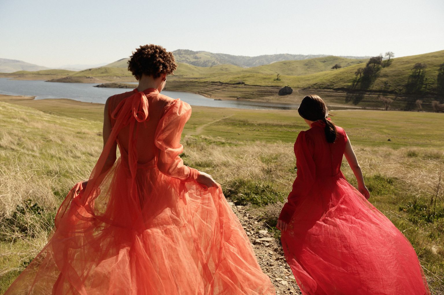 Two women in red and orange gowns walk across a grassy hill towards a blue lake on a sunny day in a landscape orientation.