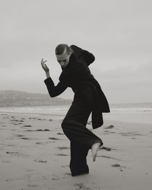 Monochrome fashion editorial image of a barefoot woman dancing on a sandy beach wearing a dark coat and trousers