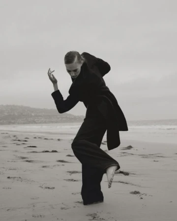 Monochrome fashion editorial image of a barefoot woman dancing on a sandy beach wearing a dark coat and trousers