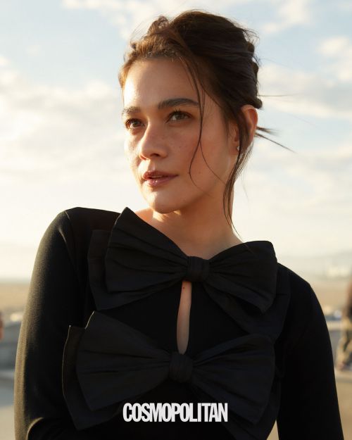 Portrait of a woman with dark hair wearing a black dress with bows, against a pale sky, featuring the Cosmopolitan logo.