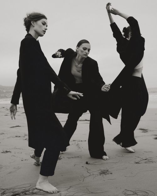 Three women in black coats and pants pose barefoot on a sandy beach under an overcast sky. Black and white fashion photography.