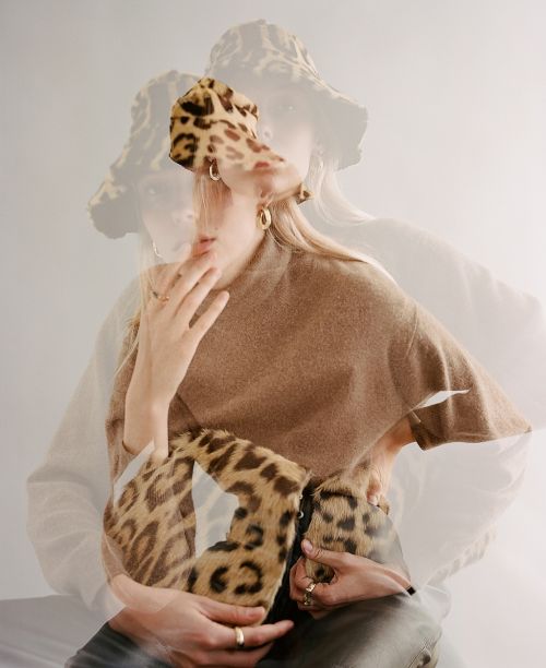 Woman poses with double exposure effect wearing a leopard print hat and handbag, gold hoop earrings, a brown top, and rings.