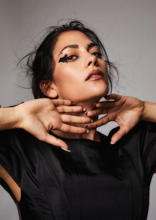 Portrait of an adult female with dark hair and dramatic black makeup, posing with her hands near her neck in a photo studio.