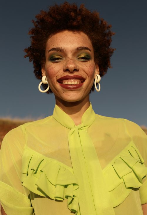A portrait of a smiling adult woman with an afro hairstyle, wearing a ruffled green top and large white hoop earrings against a blue sky.