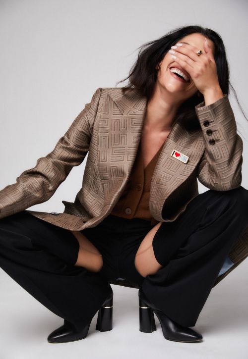 Smiling woman in blazer and heels squatting in a photo studio, wearing black pants with knee cut-outs, posing and laughing.