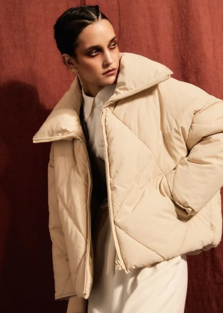 Fashion shot of a stylish woman in a cream puffer jacket against a textured reddish-brown backdrop, showcasing contemporary style.