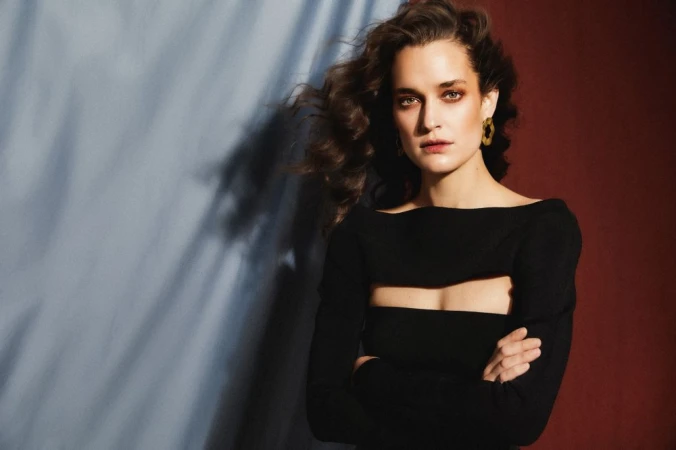Woman in a black cutout long-sleeve top wearing gold earrings against a duotone blue and deep red backdrop