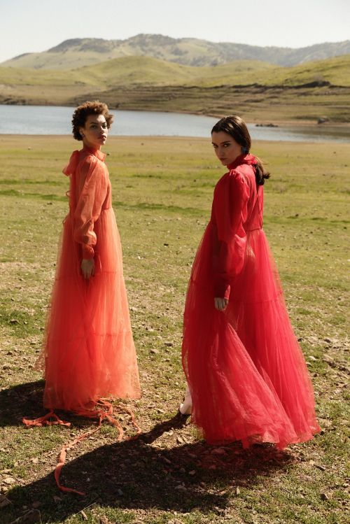 Two women in long sheer red dresses standing outdoors on a grassy hillside near a lake styled by Karlee Parrish.