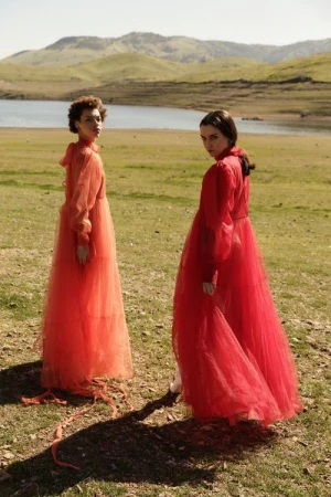 Two women in long sheer red dresses standing outdoors on a grassy hillside near a lake styled by Karlee Parrish.