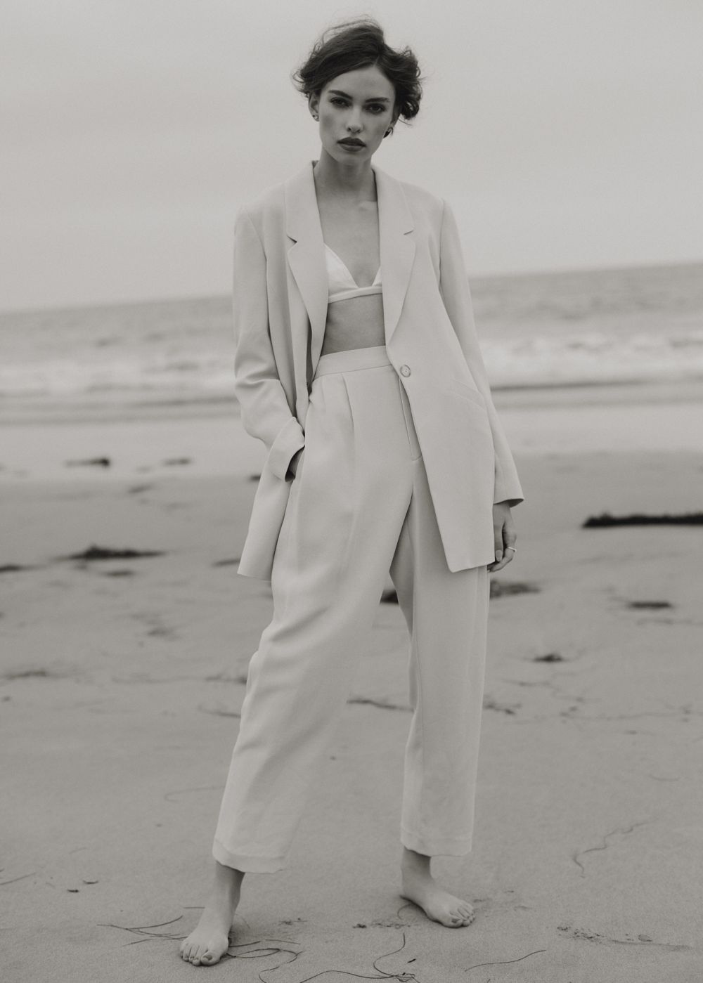 Woman poses in a linen suit on a beach, wearing a bra top and standing barefoot on the sand with the ocean in the background.