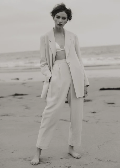 Woman poses in a linen suit on a beach, wearing a bra top and standing barefoot on the sand with the ocean in the background.