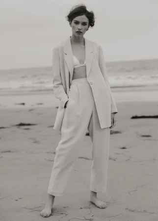 Woman poses in a linen suit on a beach, wearing a bra top and standing barefoot on the sand with the ocean in the background.