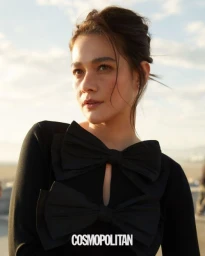 Close-up portrait of a woman wearing a black top with large bow accents against an outdoor sunset sky background.