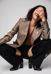 Smiling woman in patterned blazer and black trousers squatting against a plain white studio background