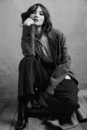 Black and white portrait of a woman wearing a plaid blazer and pinstripe trousers seated indoors.