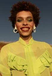 Close-up portrait of a Black woman with short curly hair wearing a lime green ruffled top and white hoop earrings against a clear blue sky