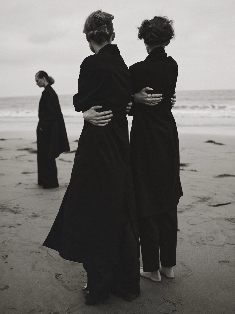 Three models in black coats standing on a sandy beach by the ocean under an overcast sky.