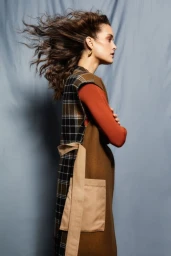 Profile portrait of a woman with long dark wavy hair blowing in the wind, wearing a plaid tunic and rust-colored top.