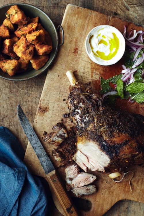 Overhead shot of a cooked lamb leg with mint and onion salad and yogurt sauce alongside fried potato cubes on a wooden surface.